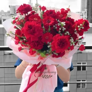 Bó hoa sinh nhật tặng bạn gái với hoa hồng đỏ, cúc mẫu đơn đỏ, baby trắng, gói bằng giấy hồng pastel, nơ đỏ, chụp trong bối cảnh ngoài trời.