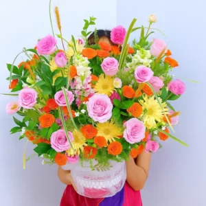 Shop hoa tươi giao tận nơi – Người cầm lẵng hoa rực rỡ gồm hồng phấn, cúc cam và lá xanh